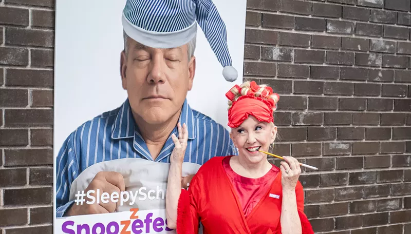 Image: a woman stands by a poster of a sleeping david schweikert