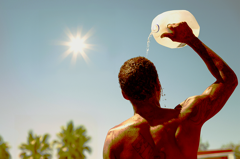 Image: A man under a glaring sun pours water over his head out of a jug.