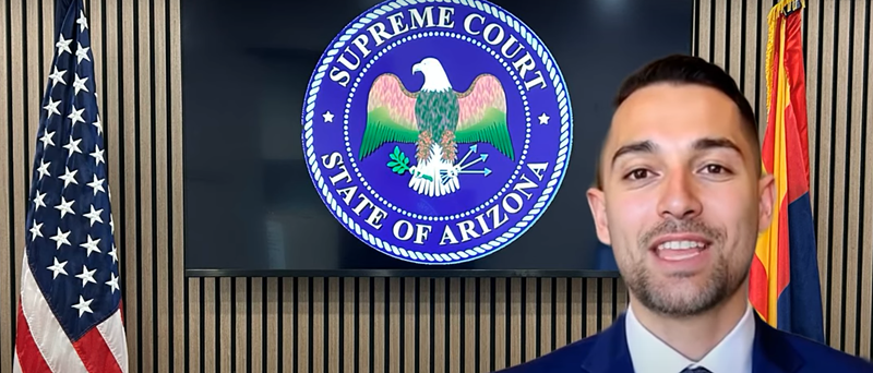 Image: an ai-generated man in a suit speaks to the camera in a "room" with the U.S. and Arizona flags and a TV with the Arizona Supreme Court seal on it