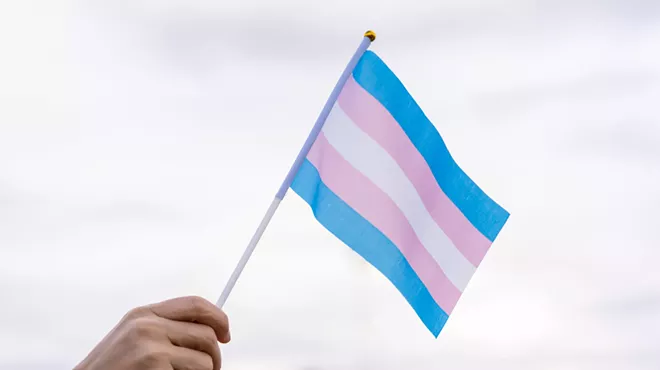 a hand waves a small flag with trans advocacy colors (pink, white, powder plue)
