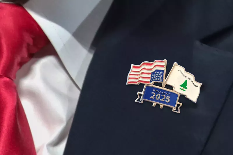 Image: A lapel pin with an upside-down U.S. flag and a flag with a pine tree that says "An Appeal To Heaven," both of which sit atop a banner saying "Project 2025"