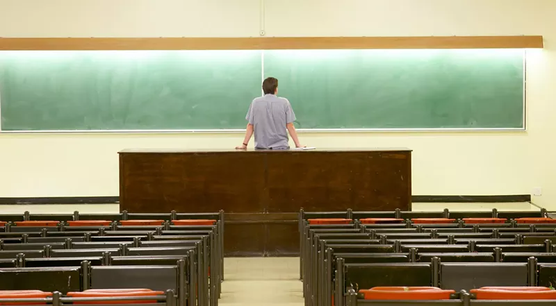 Image: a stock photo of a professor staring at an empty chalkboard in an empty classroom
