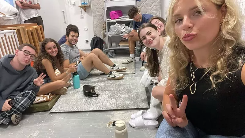 Image: young adults sit on the floor and pose for a selfie in a bomb shelter