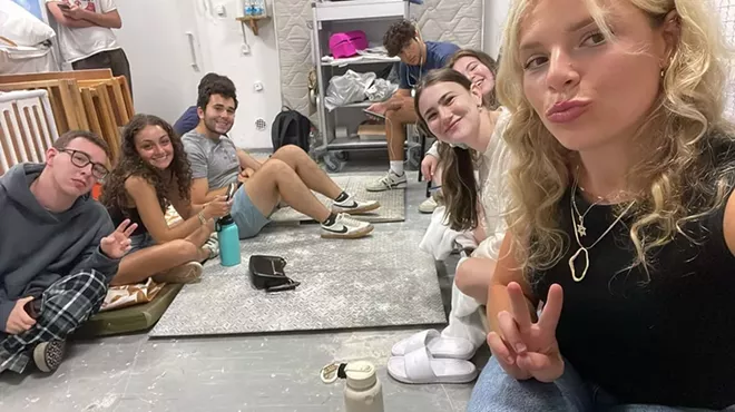 young adults sit on the floor and pose for a selfie in a bomb shelter