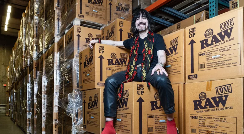 Image: a man in a black shirt and red scarf sits on a pile of boxes