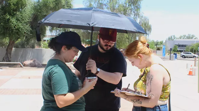 a woman watches another woman sign a form as a man holds an umbrella over both on a sunny day