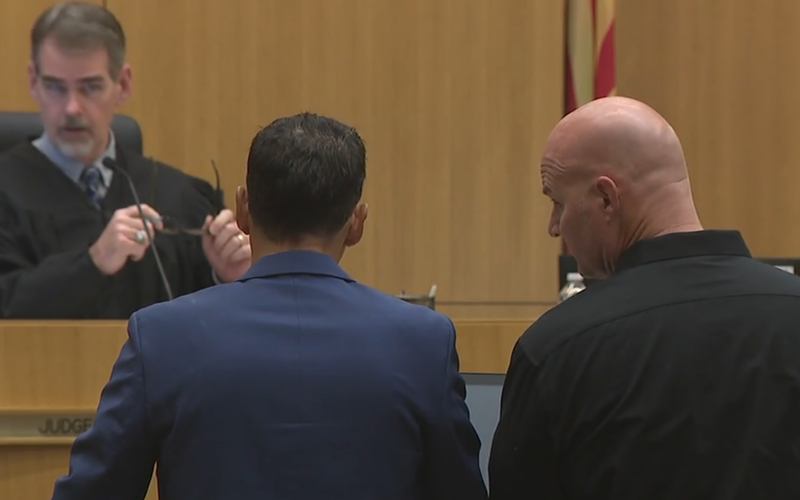 Image: a man in black in a courtroom next to a man in a suit, both looking at a judge