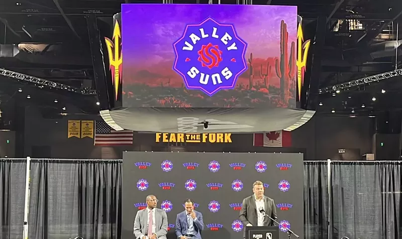 Image: A man speaking at a podium on a stage. Above him is a video board showing the logo for the Valley Suns of the G League.