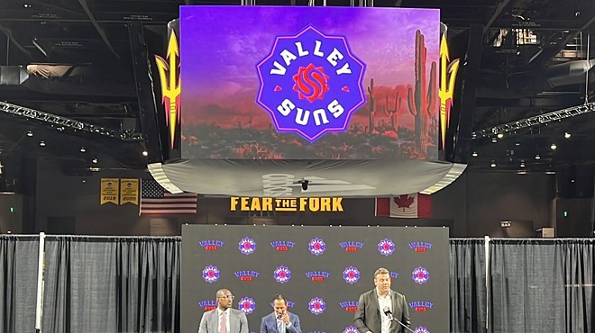 A man speaking at a podium on a stage. Above him is a video board showing the logo for the Valley Suns of the G League.