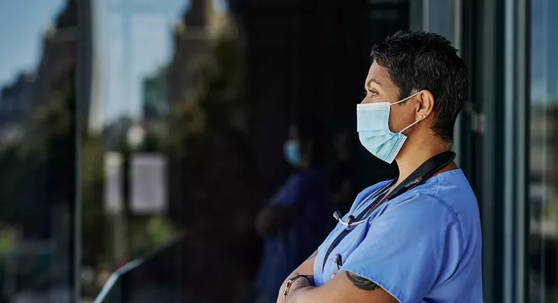 Image: a health worker in a mask stares into the distance