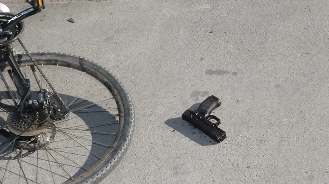 A wheel of a bicycle sits near a handgun on concrete.