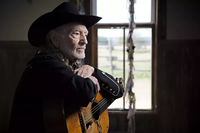 Image: Willie Nelson sits in a room holding a guitar.