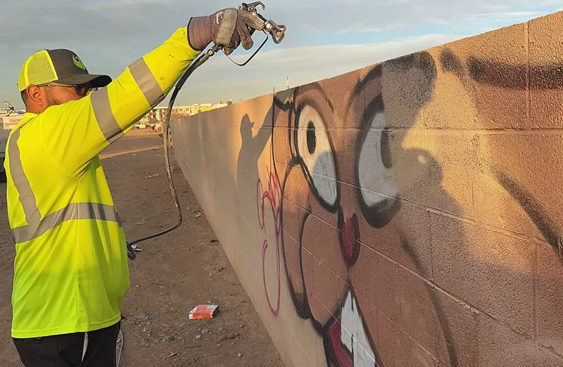 Image: a man sandblasting graffiti off a block wall