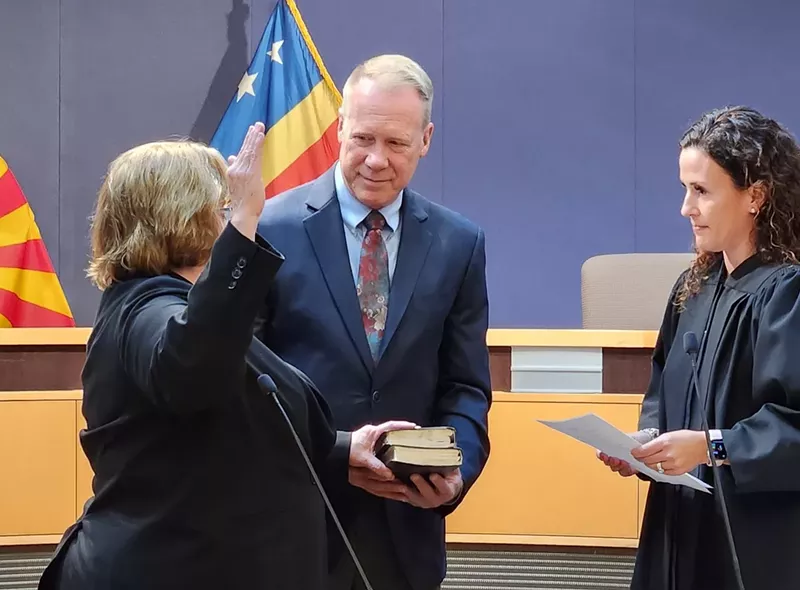 Image: A woman raising her right hand with her left on a bible in front of a female judge. A gray-haired man holds the bible