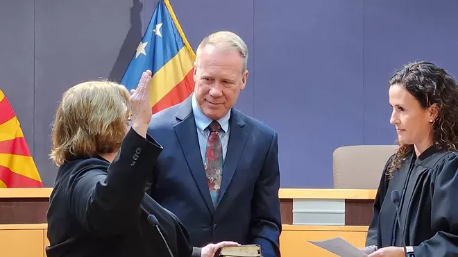 A woman raising her right hand with her left on a bible in front of a female judge. A gray-haired man holds the bible