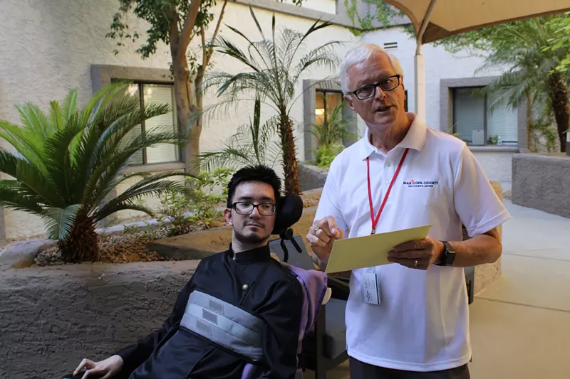 Image: a young man in a wheelchair listens as an older man in a while shirt talks