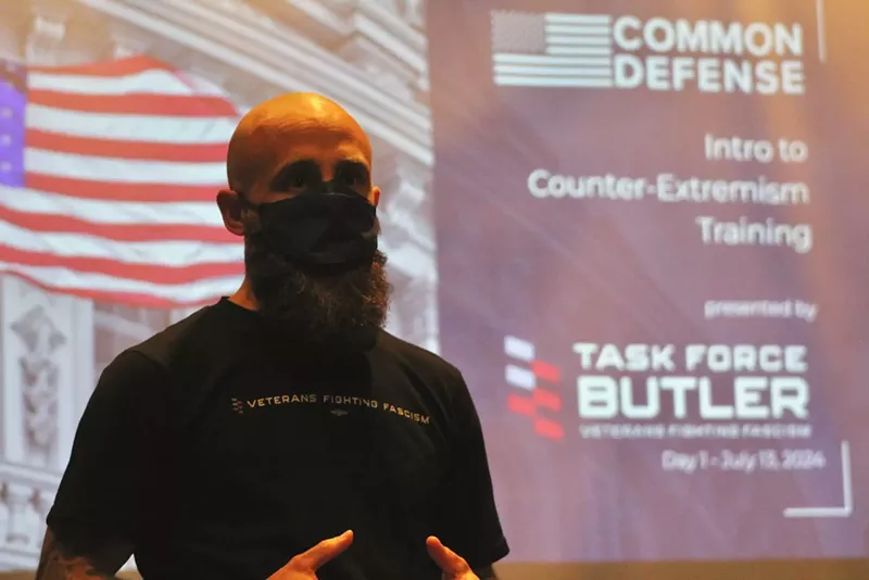 Image: A man in a face mask speaking in front of a screen
