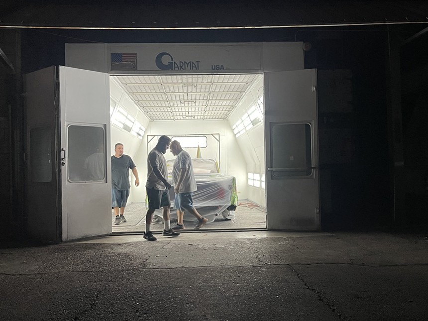 Men walking in front of a paint booth that contains a car.
