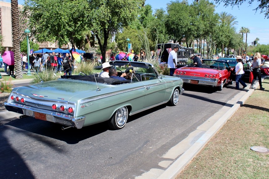 A lowrider car drives down a small boulevard with trees in the background.
