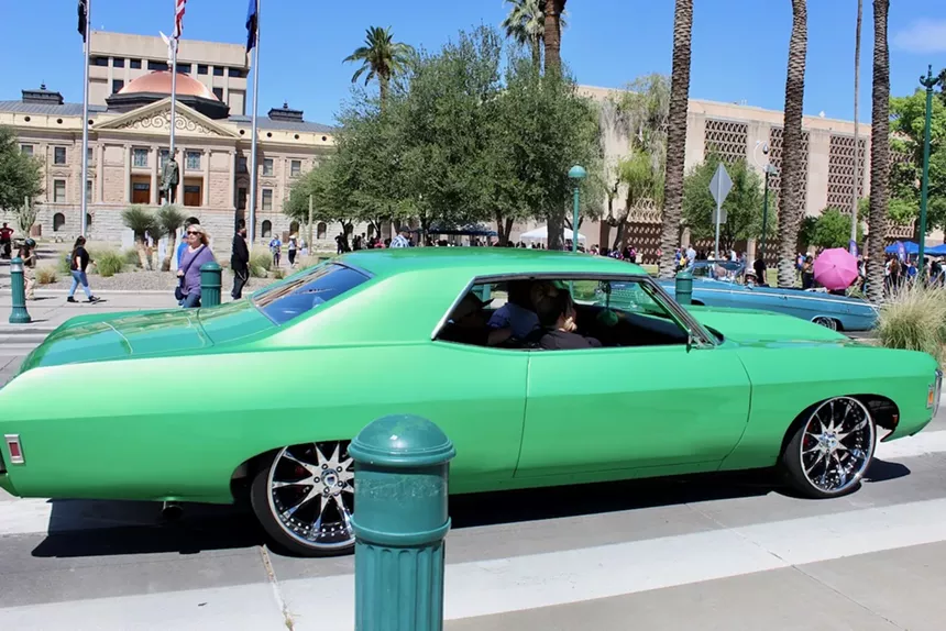 A green car in a street with a tree and buildings in the background.