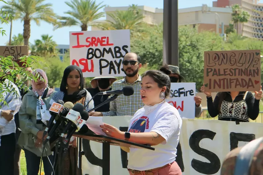 A person stands at a podium with people holding signs behind them.
