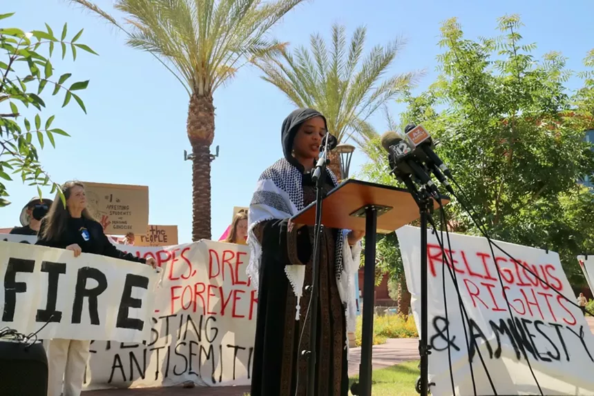 A woman speaks at a microphone stand with signs and trees in the background.