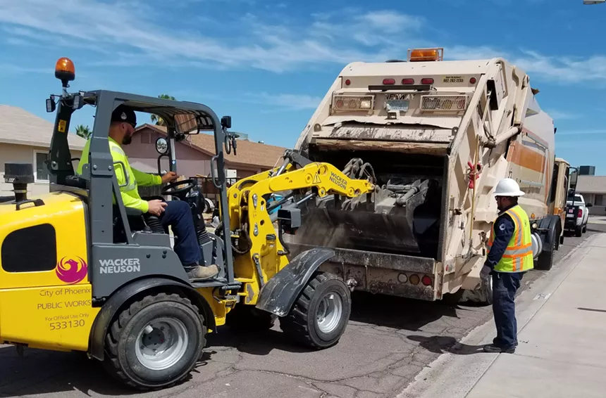 A backhoe dumping heavy trash into the back of a garbage truck.