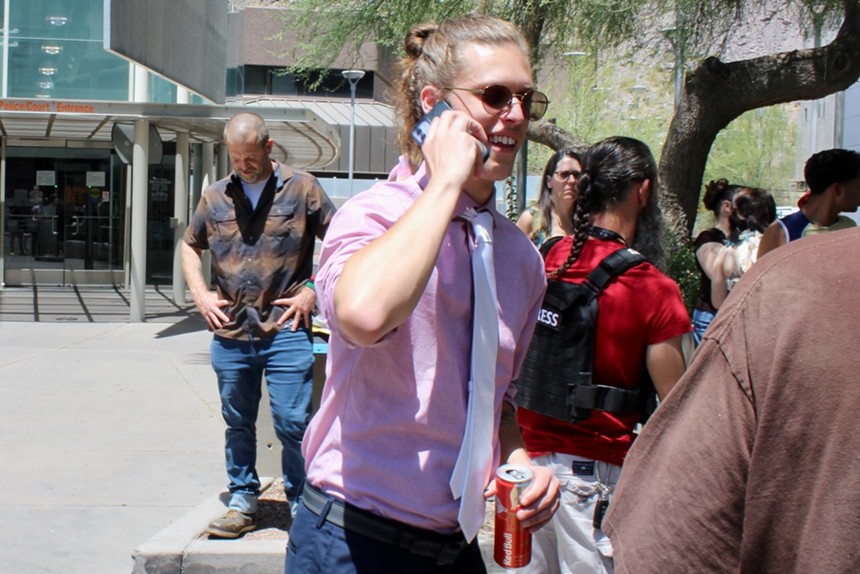 A man in sunglasses, a tie and purple shirt with rolled-up sleeves speaks on the phone.