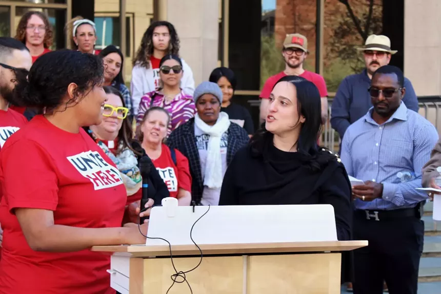 A woman at a podium looks at someone in a UNITE HERE shirt. Behind her are several supporters.