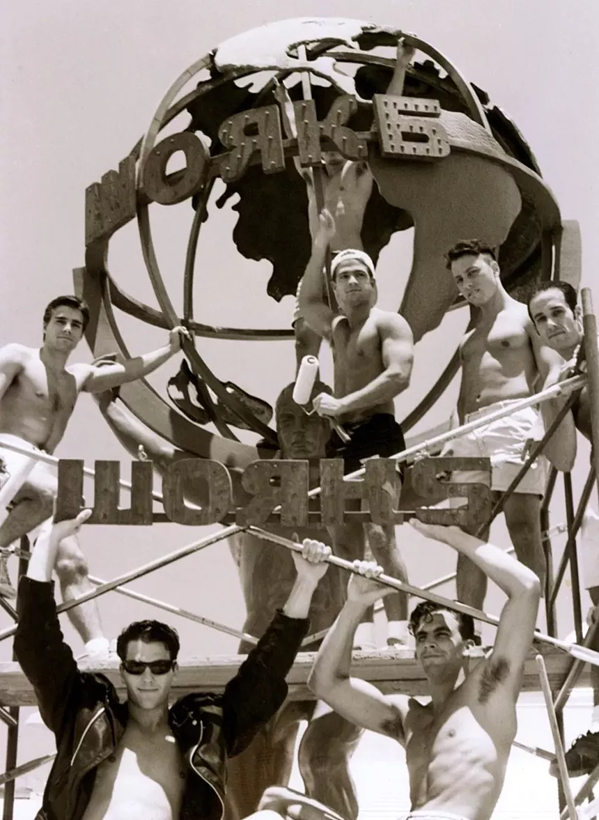 Shirtless men stand in from a metal globe of the world.