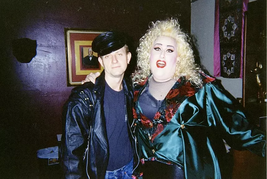 A man in a military-style cap and black leather jacket standing next to a drag queen.