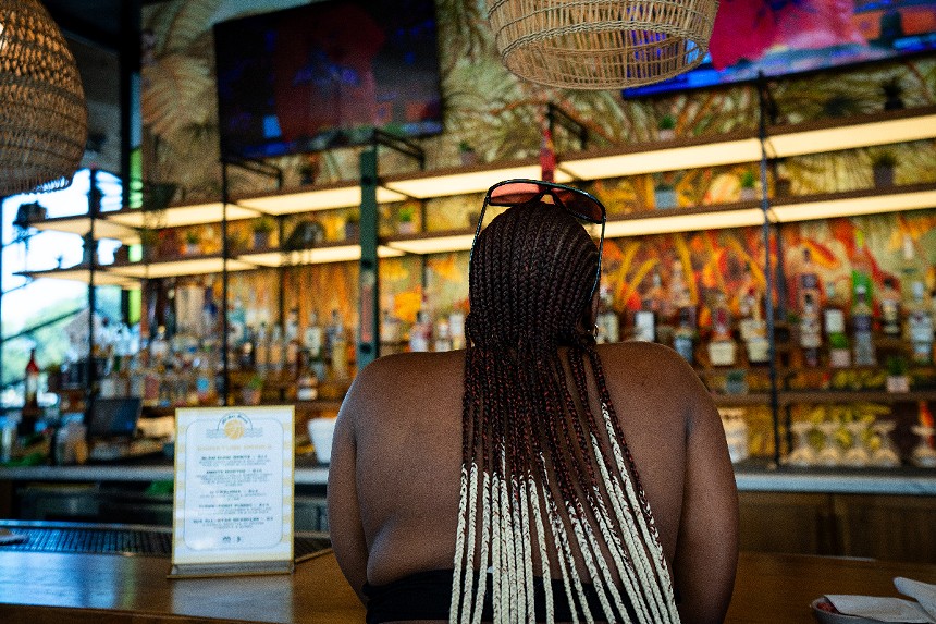 A woman sits at a bar, seen from behind, with long brown and white braids