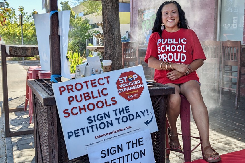 a woman in a "public school proud" shirt sits next to a sign that says "Protect Public Schools. Sign the Petition Today. Stop Voucher Expansion."