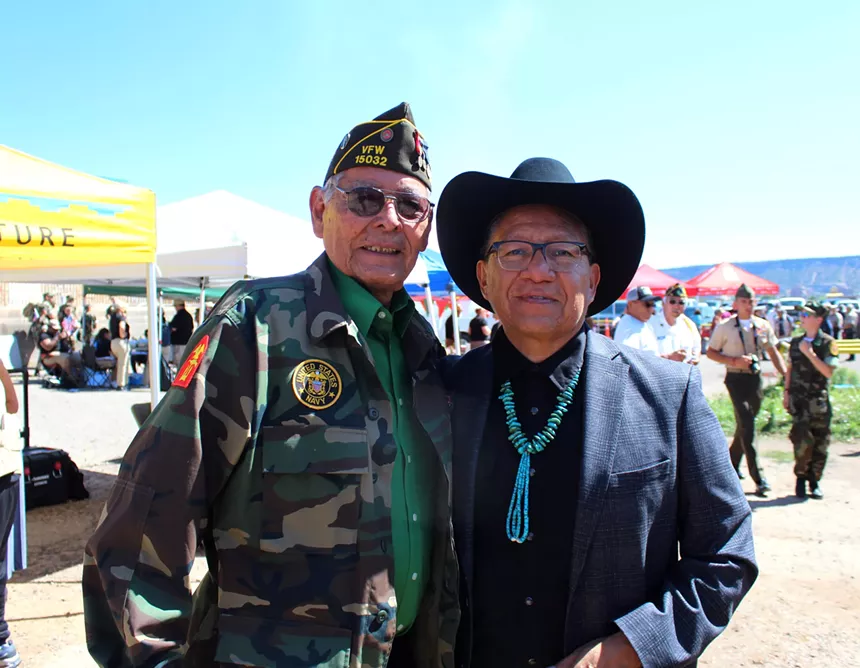 Two men stand smiling and facing forward while putting arms around each other. The sky behind is bright and blue.