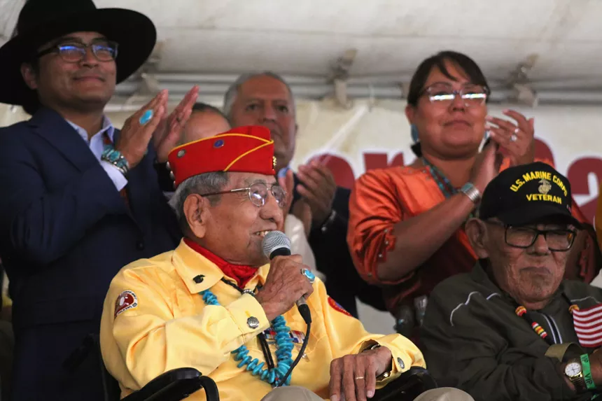 A man holds a microphone and speaks while sitting with people standing behind him.