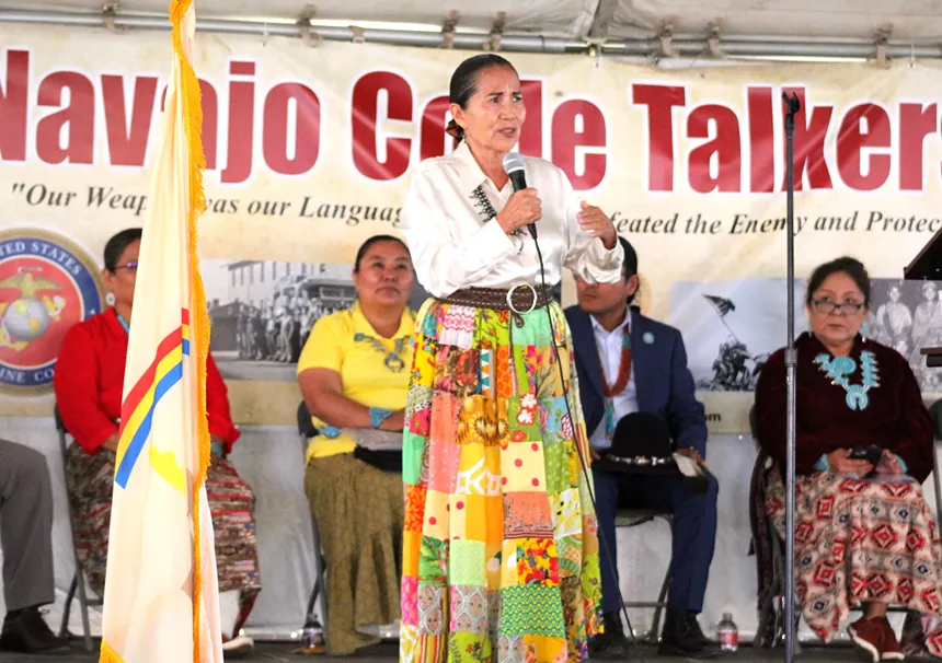 A woman holds a microphone and speaks with people sitting behind her.