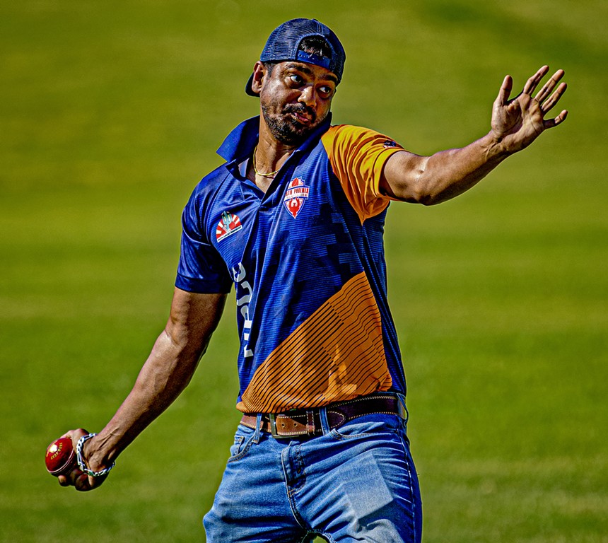 a man in an orange and blue shirt and jeans bowls a cricket ball