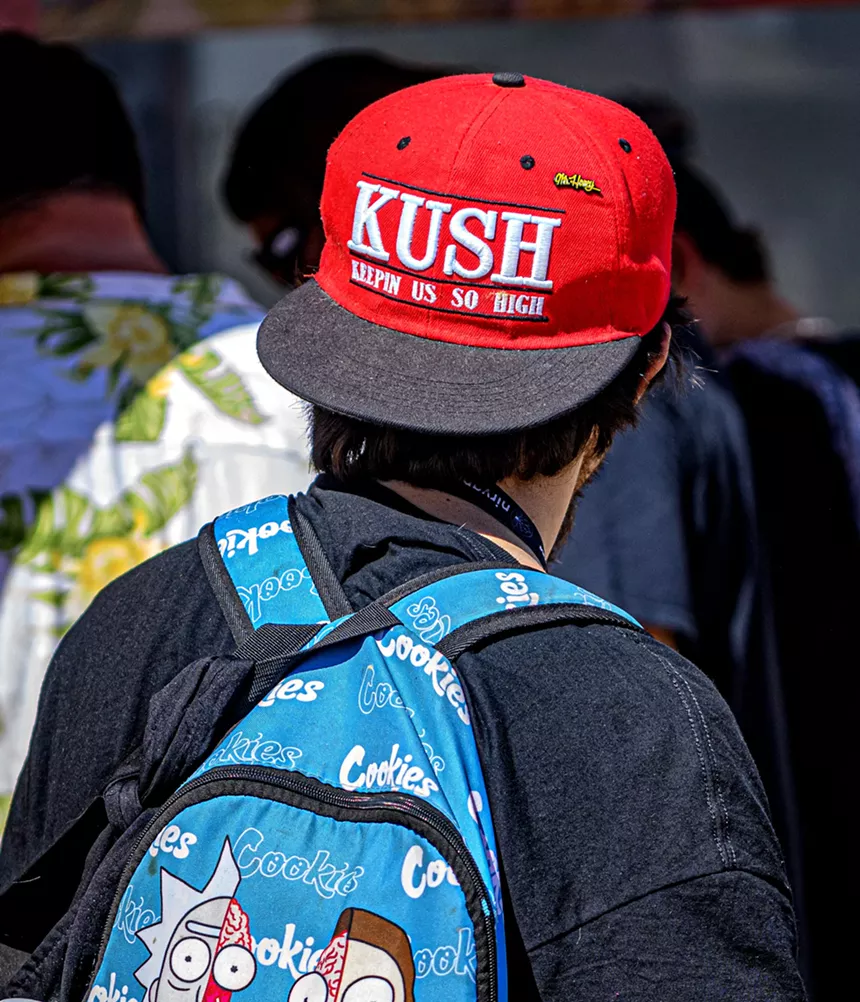 seen from behind, a man in a hat that says "KUSH: Keepin Us So High" and wearing a Rick & Morty backpack.