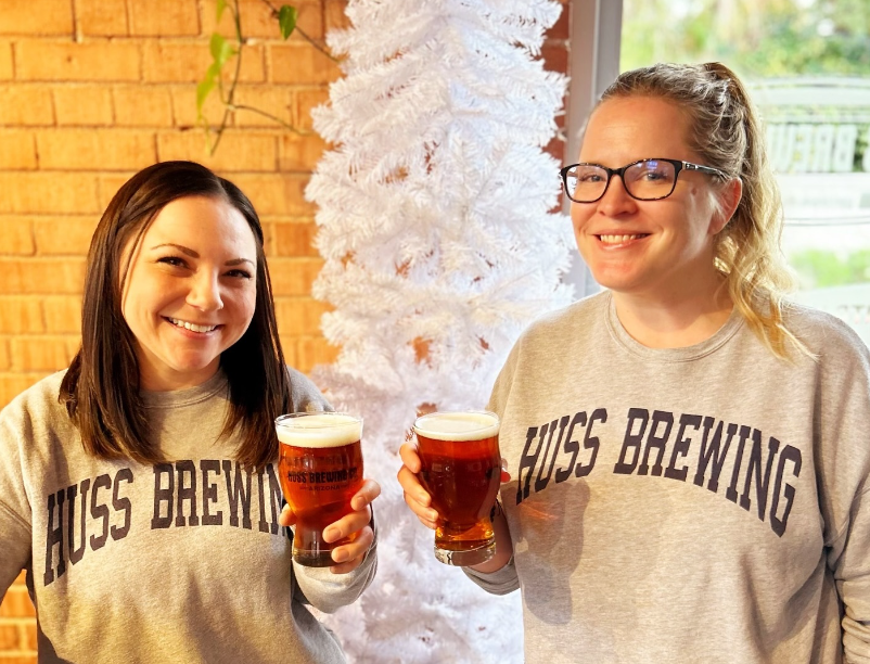Two women in matching Huss Brewing sweatshirts hold pints of the brewery's seasonal Christmas Ale.