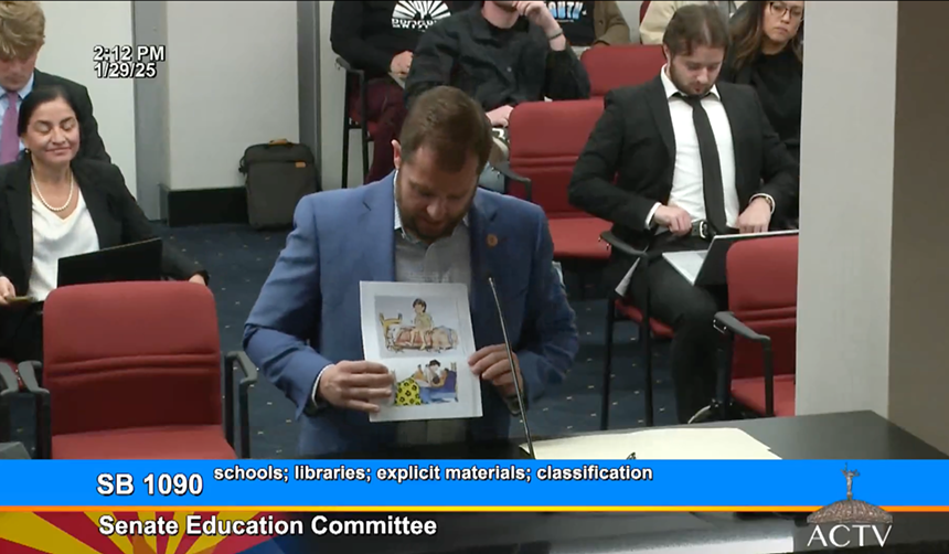 a person in a suit points to illustrations in a book in a legislative hearing