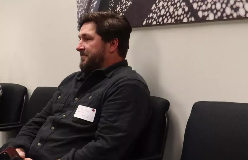 a man in a black button-down shirt with a beard sits in a chair