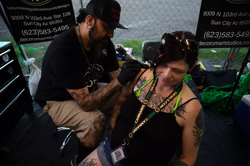 a woman sits as a tattoo artist inks her upper arm