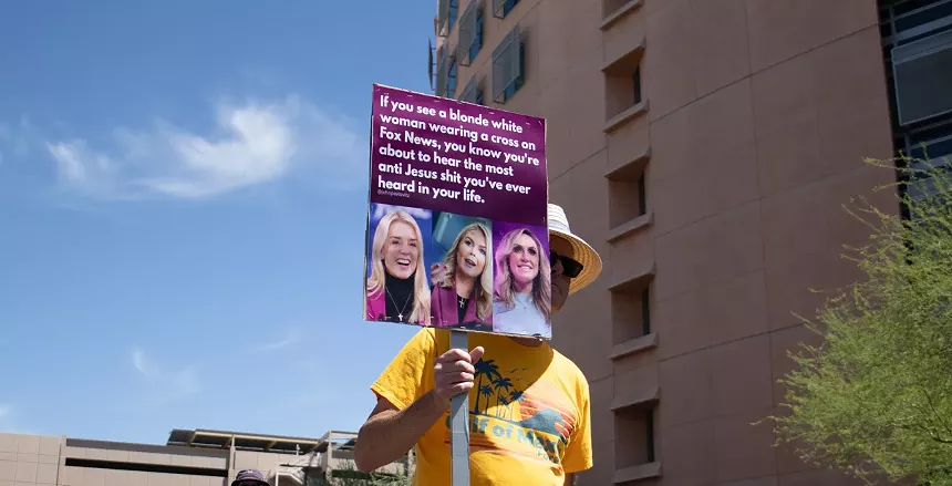 a protester holds a sign with pictures of pam bondi and karoline leavitt that says "if you see a blonde white woman wearing a cross on Fox News, you know you're about to hear the most anti jesus shit you've ever heard in your life"