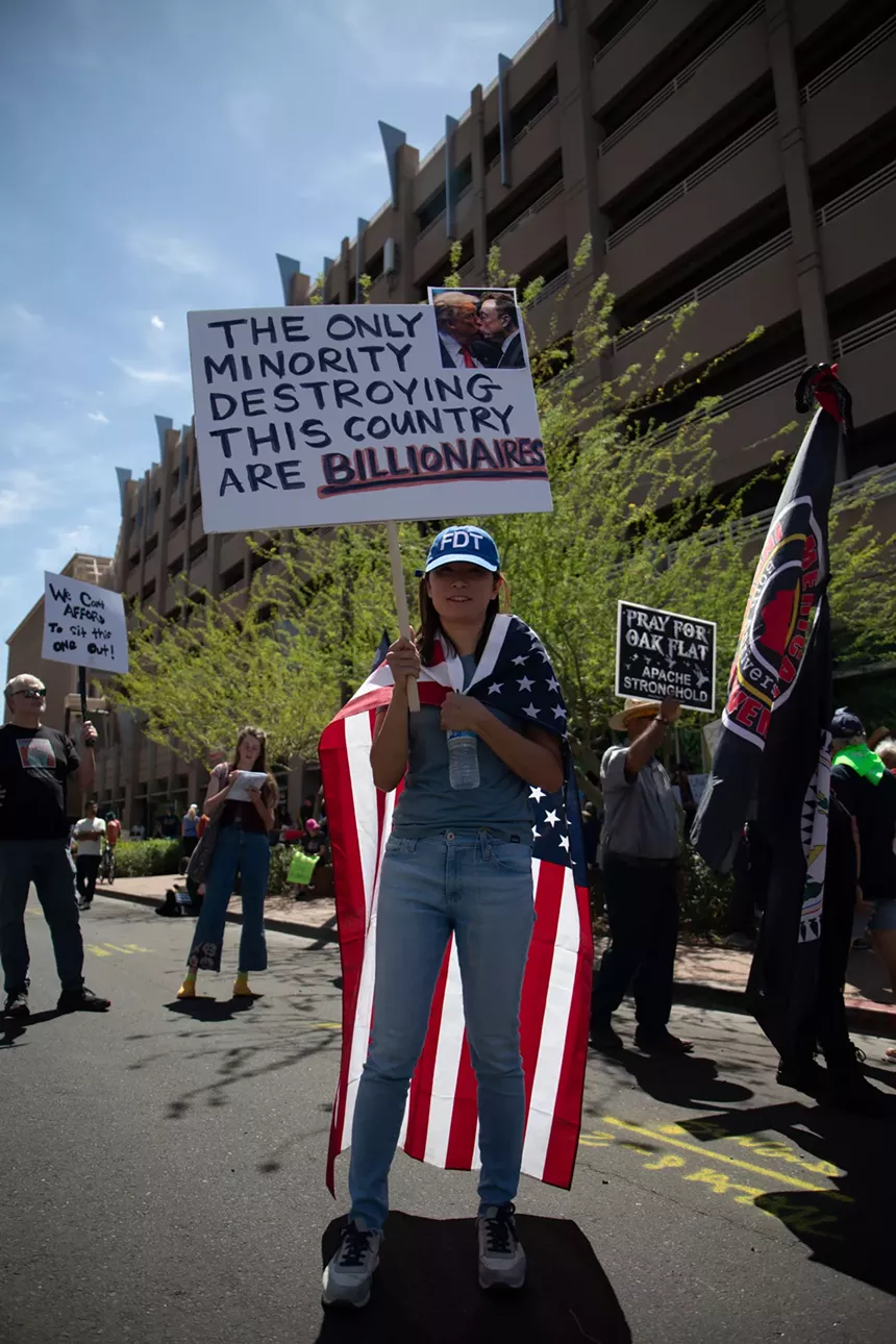 a protester draped in an american flag holds a sign that features a fake photo of Donald Trump and Elon Musk kissing