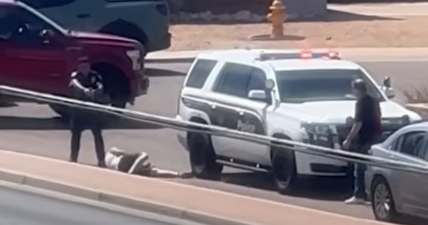 a man lies on the ground next to a police car while an officer points a gun