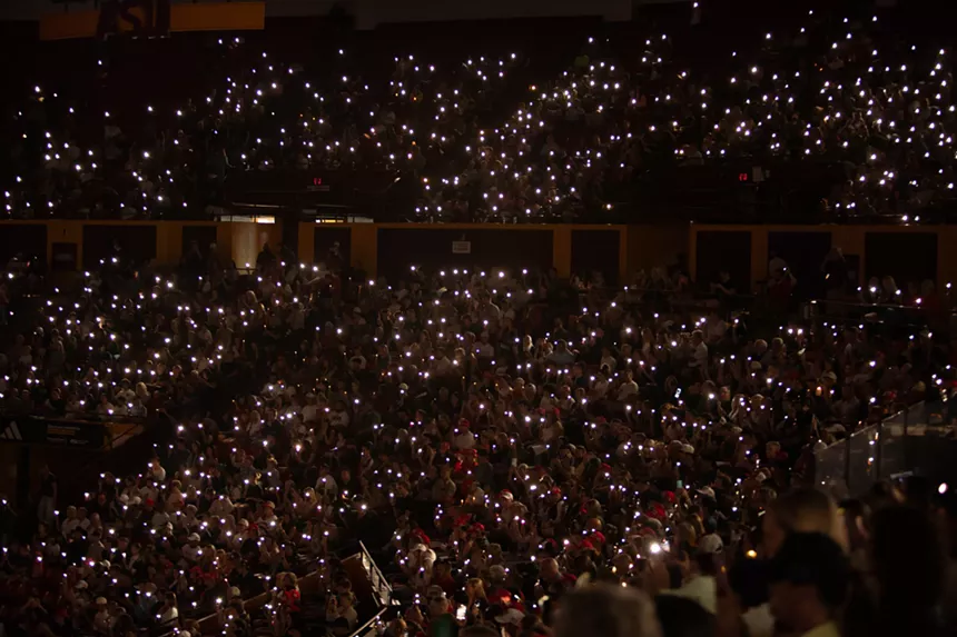 a sea of people in a dark arena hold up their phones with the flashlight on