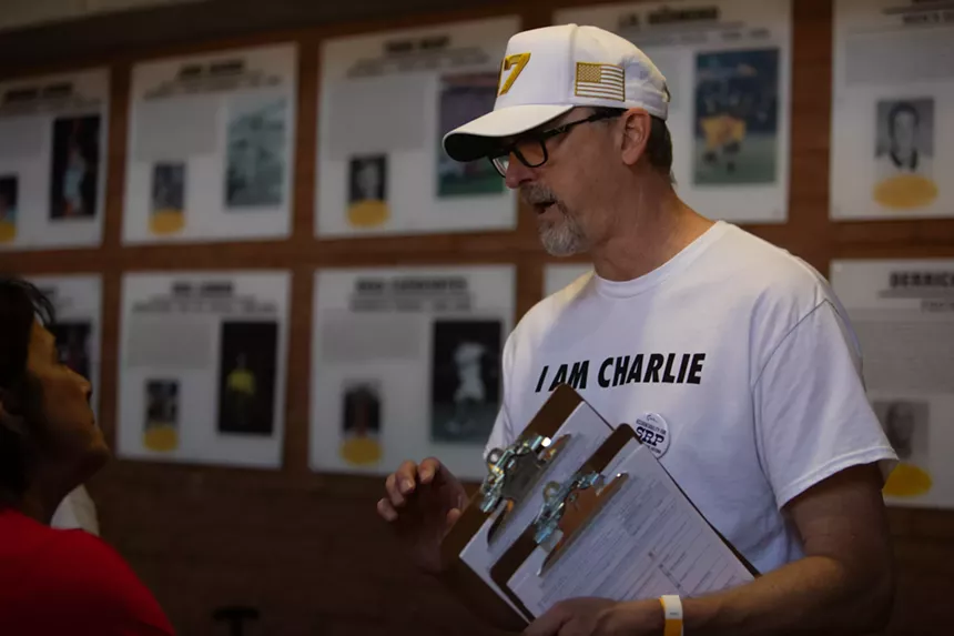 a man in a white hat and a white "i am charlie" shirt holds a clipboard while talking