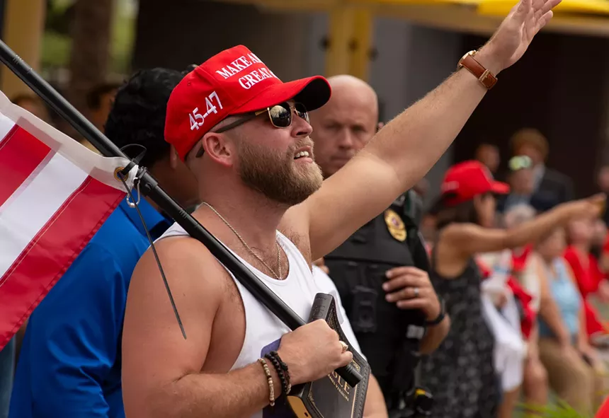 a man in a MAGA hat holds a hand up in worship while his other hand holds a bible and american flag