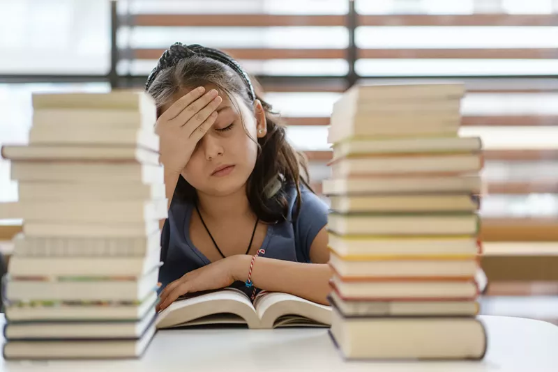 Image: an elementary school girl puts a hand on her head in exasperation while stacks of books sit in front of her
