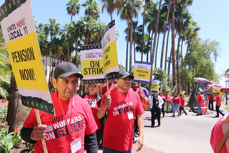 Image: Hotel workers in red shirts hold picket signs with phrases like "On Strike!" and "Pension Now!" In the background is an inflatable rat.